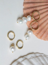 Gold hoop earrings with pearl charms on them sitting next to a peach and white seashell